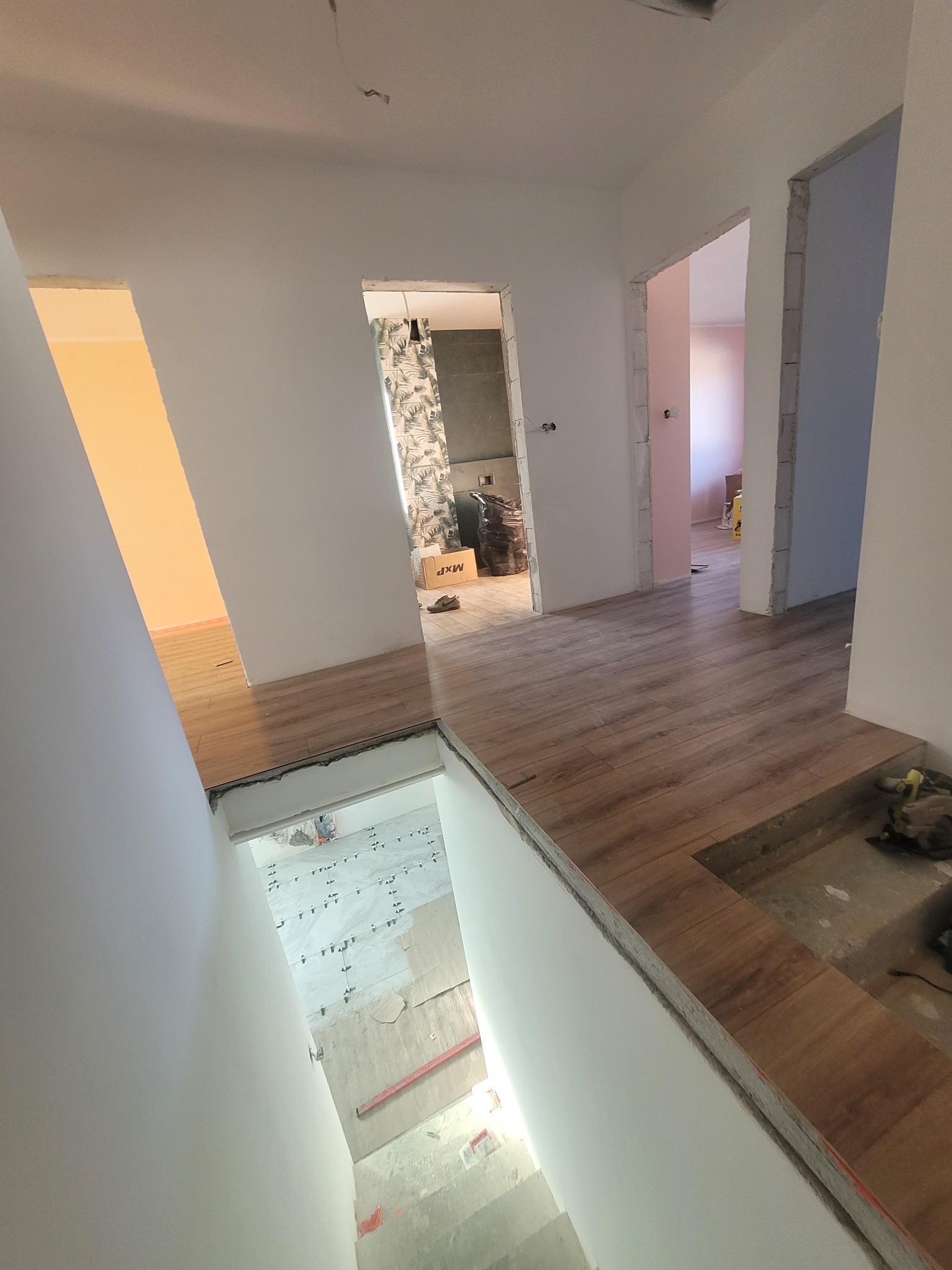 Wnętrze w trakcie prac wykończeniowych: ułożone panele podłogowe w korytarzu, widok na łazienkę z tapetą w liście i częściowo wyłożoną płytkami podłogę w innym pomieszczeniu, widoczne płytki...