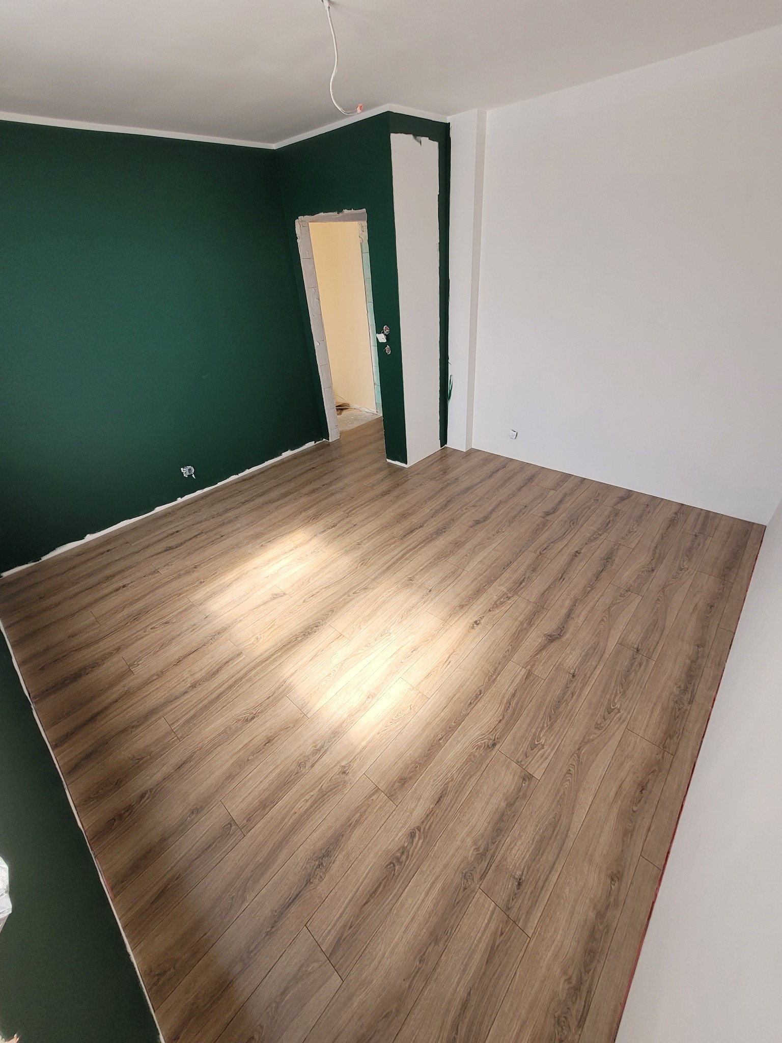Pokój po remoncie z ułożonymi panelami podłogowymi w odcieniach brązu, ściana pomalowana na ciemnozielony kolor, widoczne niedociągnięcia malarskie przy podłodze.