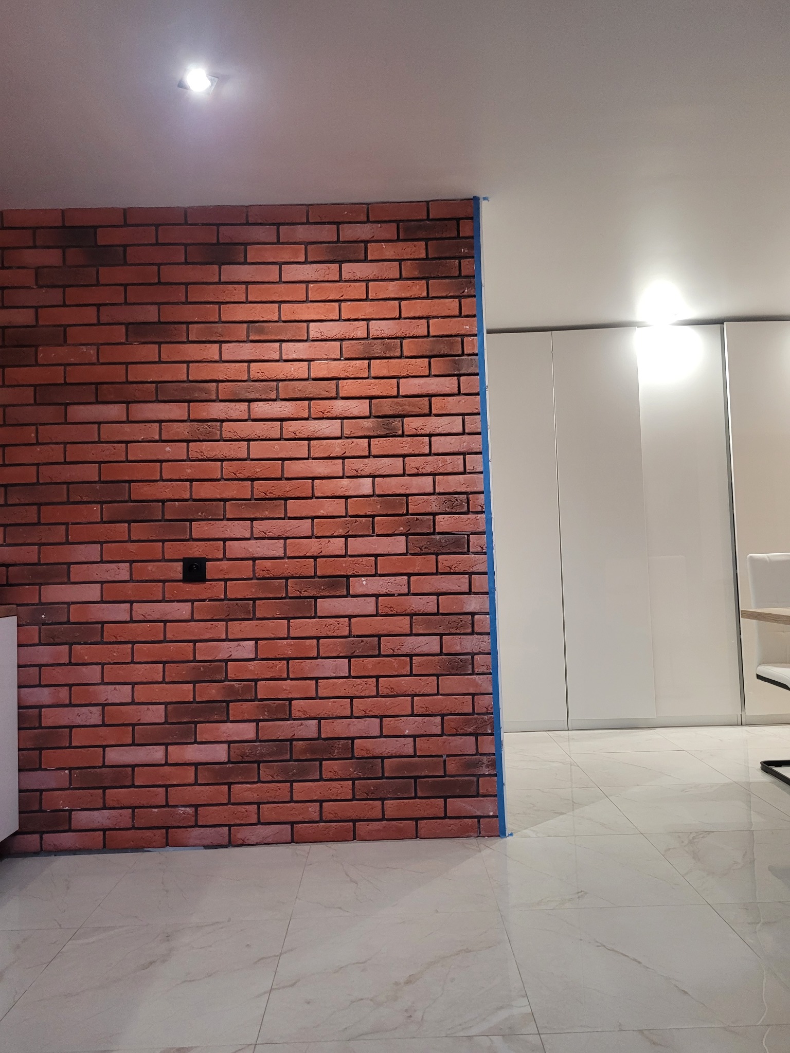 Ściana z cegieł dekoracyjnych w odcieniach czerwieni i brązu, z widocznym gniazdkiem elektrycznym, przylegająca do białej ściany z szafami wnękowymi i marmurową podłogą. Zabezpieczenie niebieską...