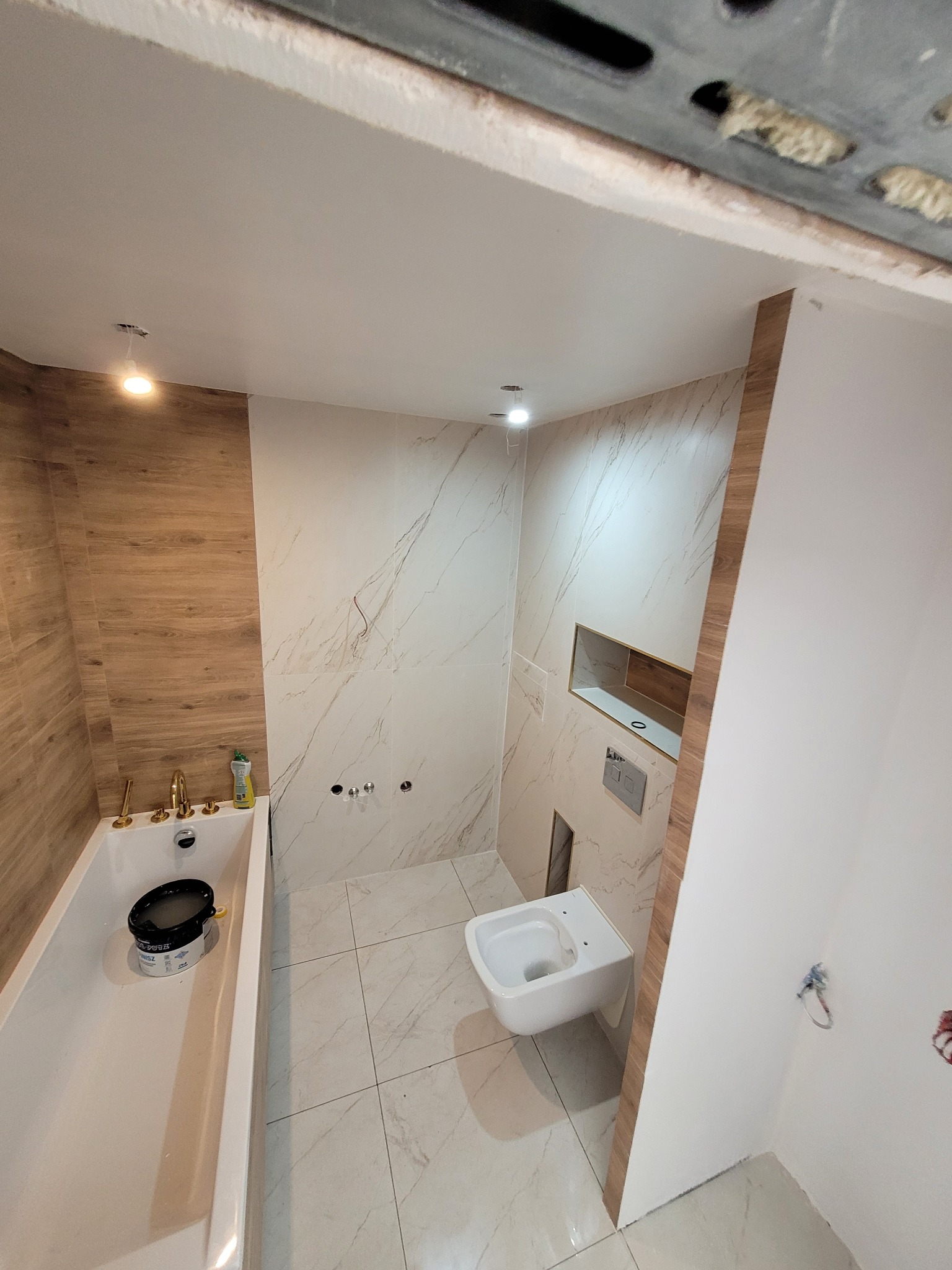 Wykończona łazienka z drewnopodobnymi płytkami na ścianie przy wannie, białymi marmurkowymi płytkami na pozostałych ścianach i podłodze, podwieszaną toaletą oraz wnęką w ścianie.