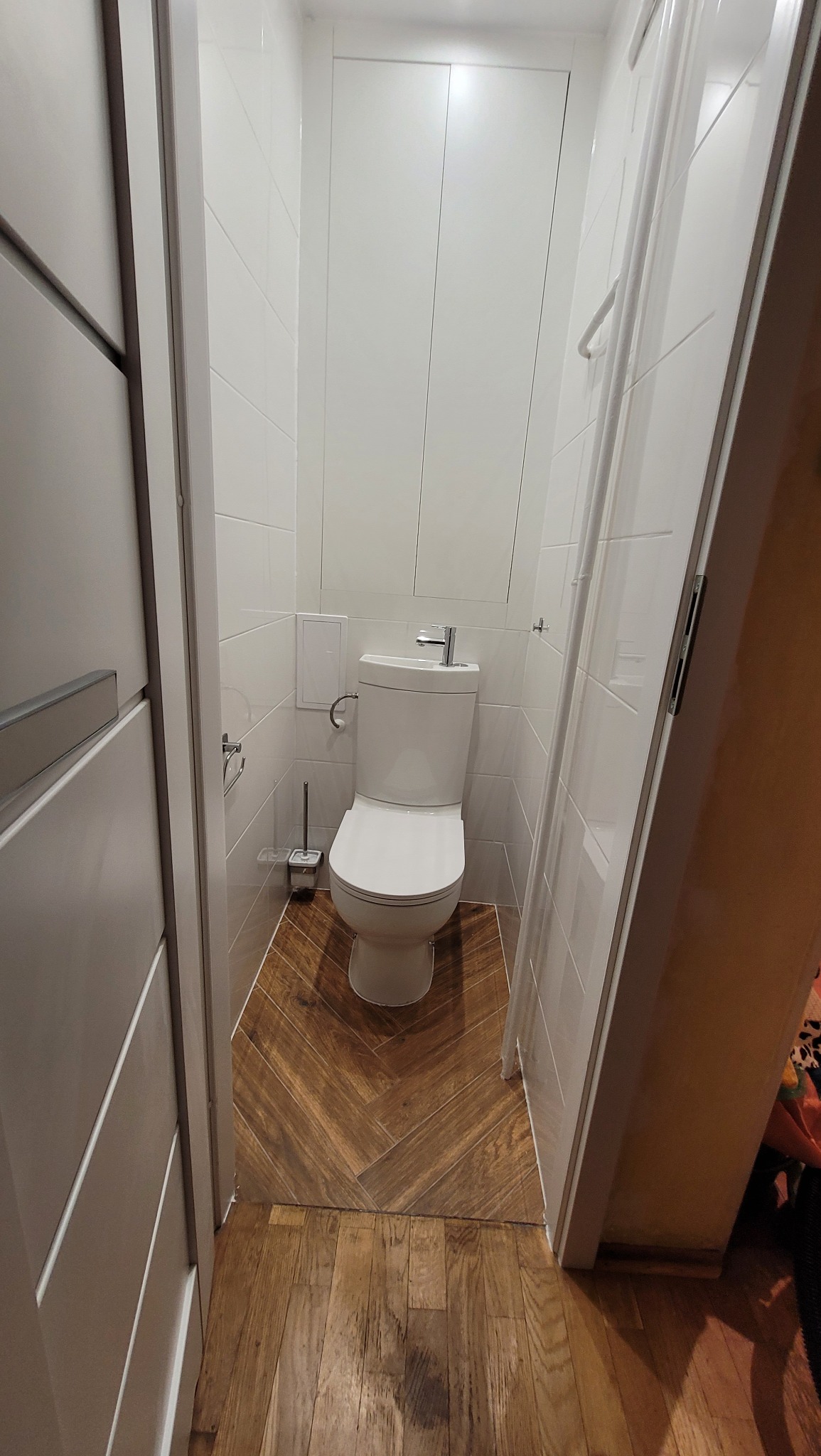 Wąska toaleta po remoncie z białą armaturą, białymi płytkami ściennymi i podłogą wyłożoną drewnopodobnymi płytkami ułożonymi w jodełkę.