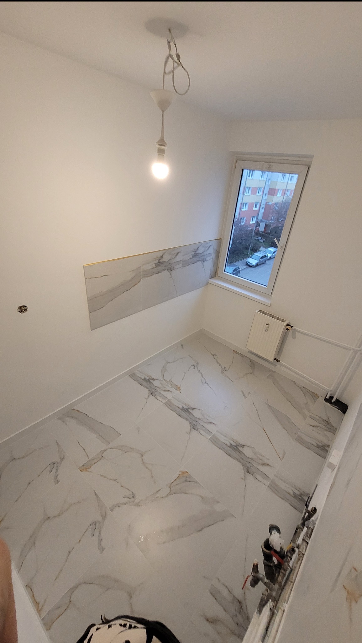 Wnętrze wykończonej łazienki z białymi płytkami imitującymi marmur na podłodze i częściowo na ścianie, widok na okno dachowe i grzejnik, w tle instalacja wodna