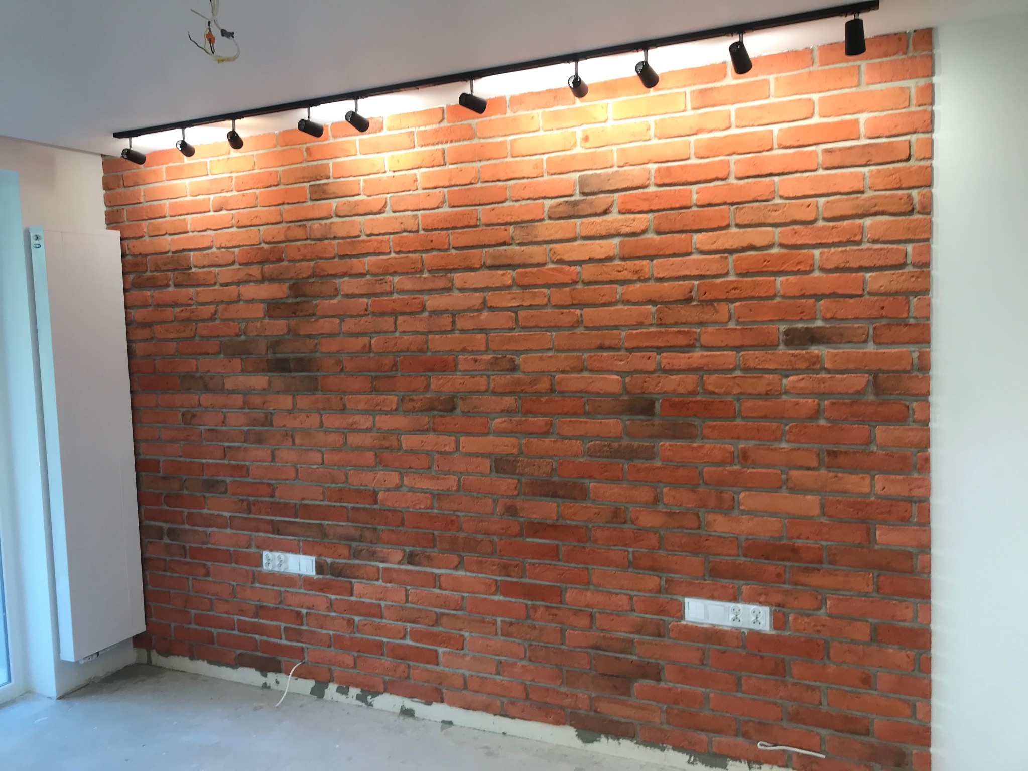 Ściana z cegły klinkierowej w pomieszczeniu z zamontowanym oświetleniem szynowym i gniazdkami elektrycznymi, widoczne przewody do podłączenia lamp.