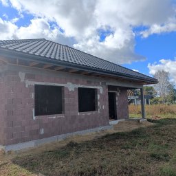 Twój Dach - Dom w trakcie budowy z ciemną dachówką na dachu i widoczną więźbą, murowany z bloczków, z otworami okiennymi i drzwiowymi, na tle błękitnego nieba z chmurami.