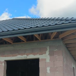 Twój Dach - Fragment nowo budowanego domu z widoczną konstrukcją dachu pokrytą ciemną dachówką ceramiczną i zamontowanymi szarymi rynnami, drewniana więźba dachowa oraz nieotynkowane ściany z pustaków...