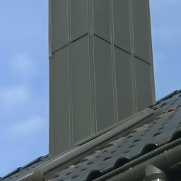 Twój Dach - Nowoczesny, grafitowy komin wentylacyjny z blachy na dachu z ciemnej dachówki ceramicznej, widoczny system rynnowy w kolorze komina, błękitne niebo w tle.