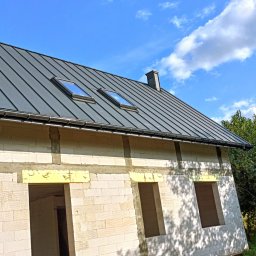 Twój Dach - Nowoczesny, grafitowy dach z dwoma oknami dachowymi na surowym budynku z białego bloczka, widok z dołu na błękitne niebo z chmurami.