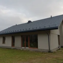 Nowoczesny dom jednorodzinny z ciemnoszarą dachówką ceramiczną i orynnowaniem w kolorze dachu, widok z boku na tle pochmurnego nieba.