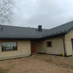 Dom jednorodzinny w trakcie wykończenia z ciemnoszarą dachówką ceramiczną, widoczne rynny, obróbki blacharskie komina i niezabudowane otoczenie.