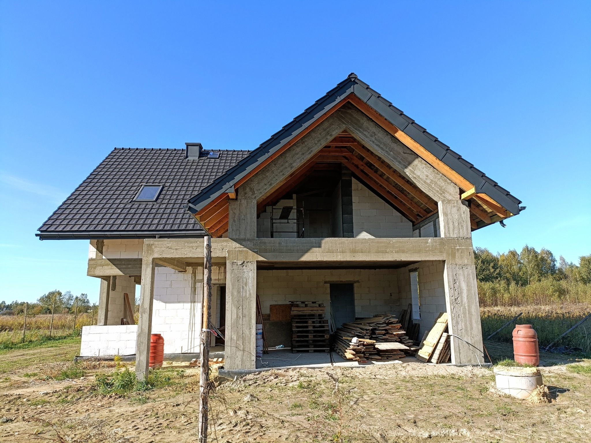 Dom w stanie surowym otwartym z częściowo wykonanym dachem z dachówki ceramicznej, widoczne elementy więźby dachowej i pustaki ceramiczne, na pierwszym planie teren budowy z materiałami.