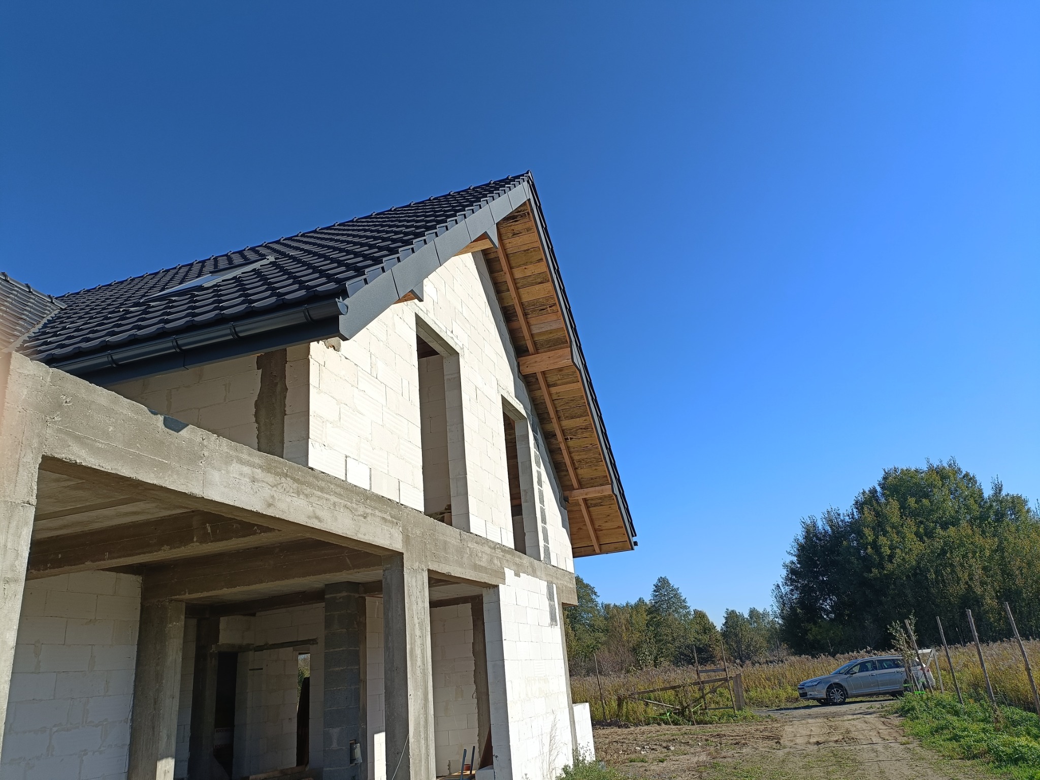 Niedokończony dom w stanie surowym otwartym z widocznym dachem pokrytym ciemną dachówką, drewnianą więźbą i betonową konstrukcją szkieletową, na tle błękitnego nieba i zieleni.