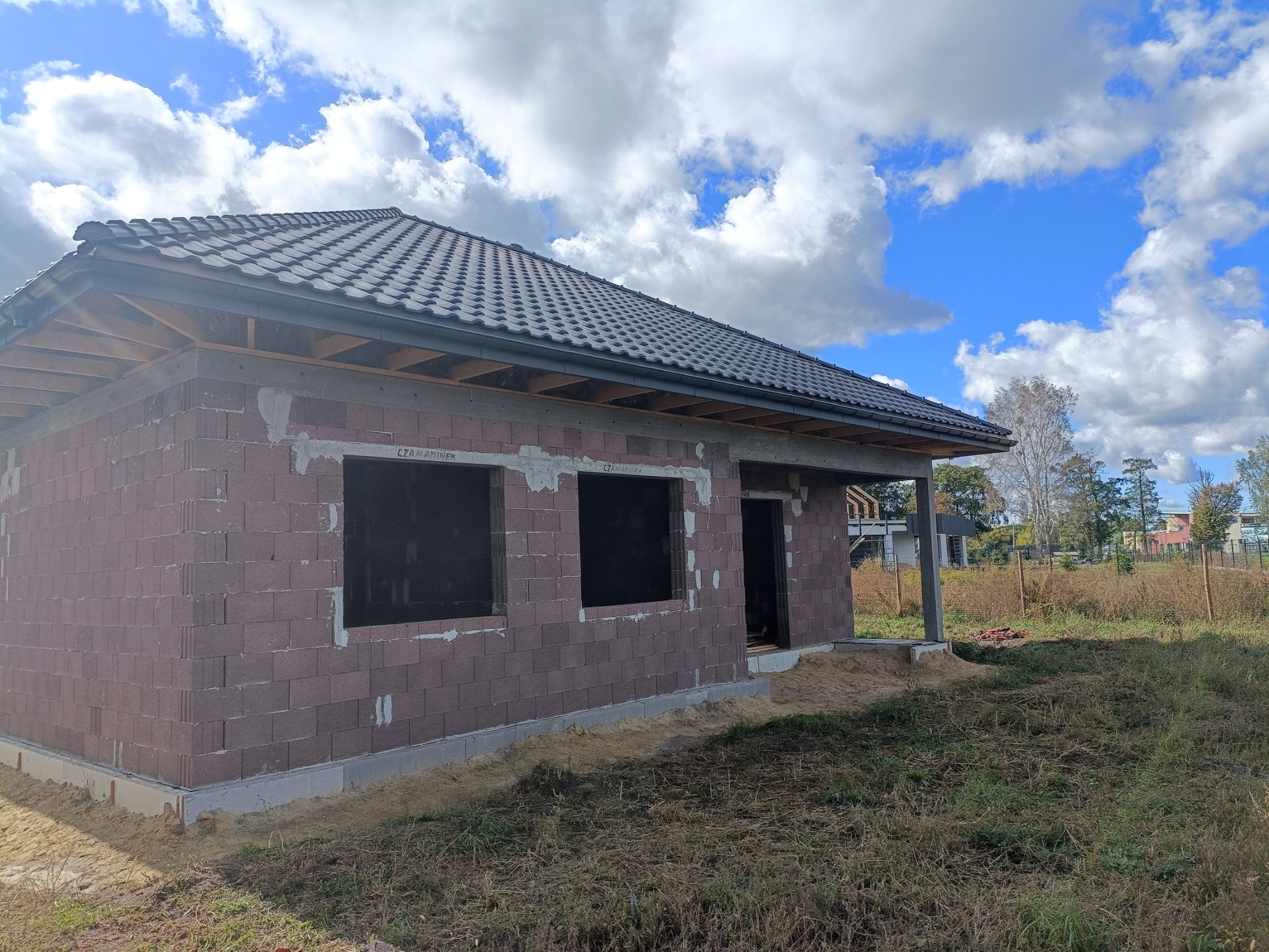 Dom w trakcie budowy z ciemną dachówką na dachu i widoczną więźbą, murowany z bloczków, z otworami okiennymi i drzwiowymi, na tle błękitnego nieba z chmurami.
