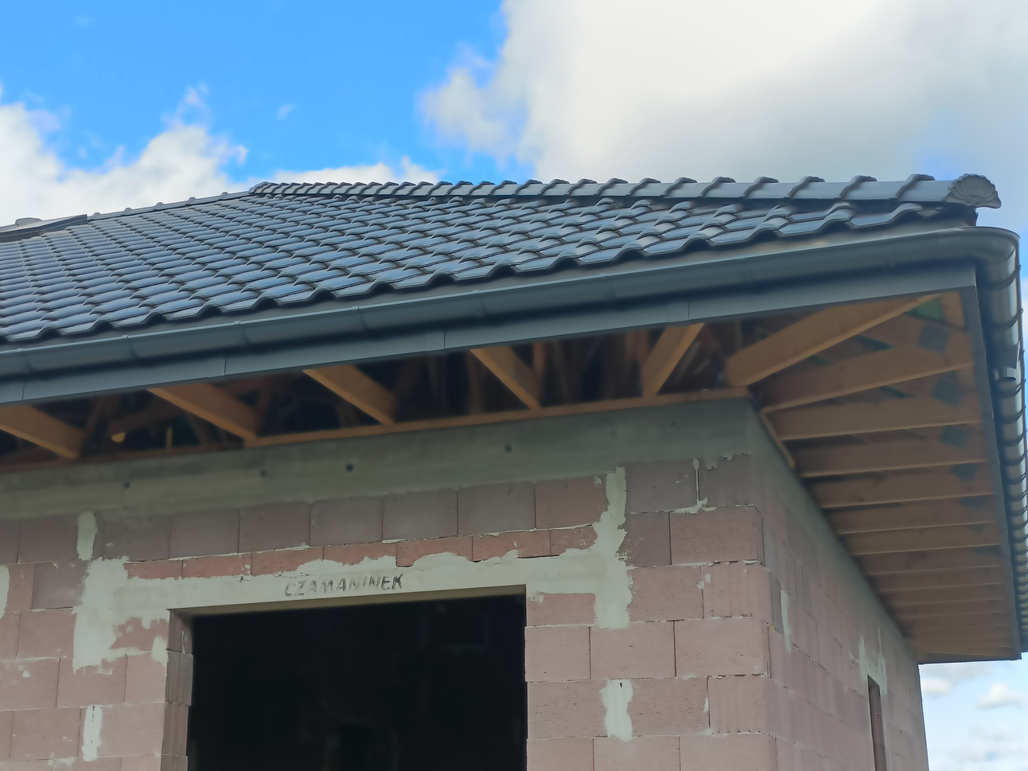 Fragment nowo budowanego domu z widoczną konstrukcją dachu pokrytą ciemną dachówką ceramiczną i zamontowanymi szarymi rynnami, drewniana więźba dachowa oraz nieotynkowane ściany z pustaków...