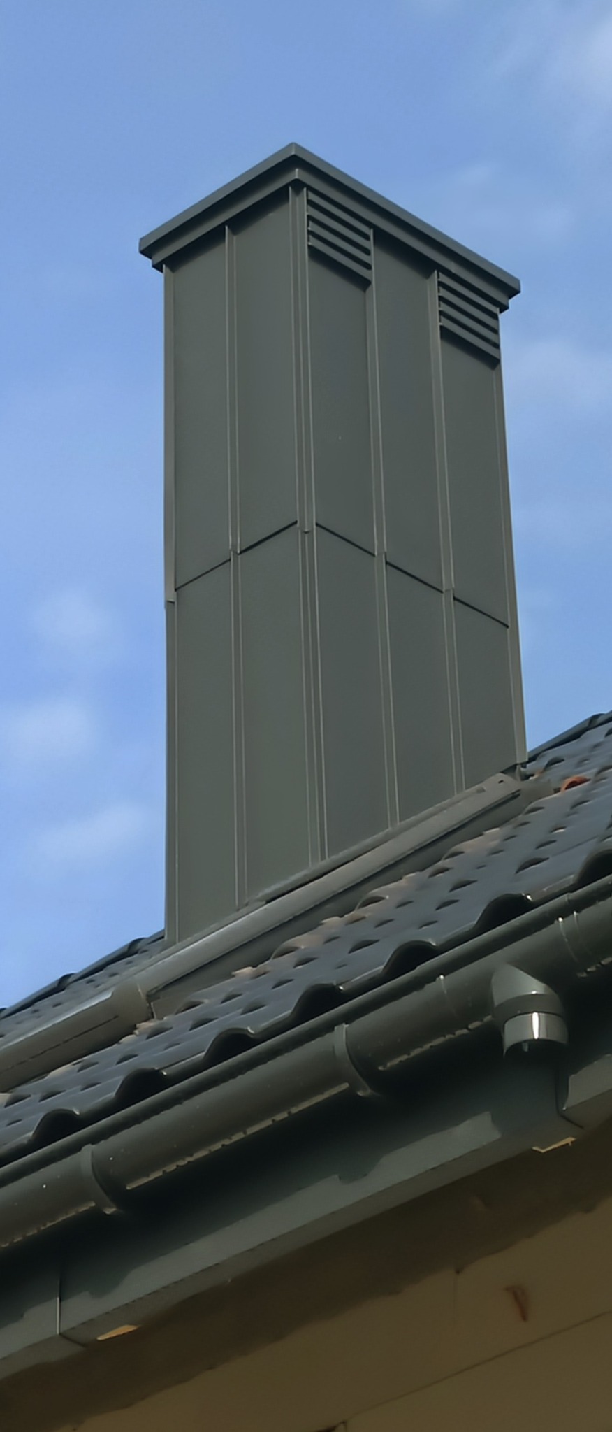 Nowoczesny, grafitowy komin wentylacyjny z blachy na dachu z ciemnej dachówki ceramicznej, widoczny system rynnowy w kolorze komina, błękitne niebo w tle.