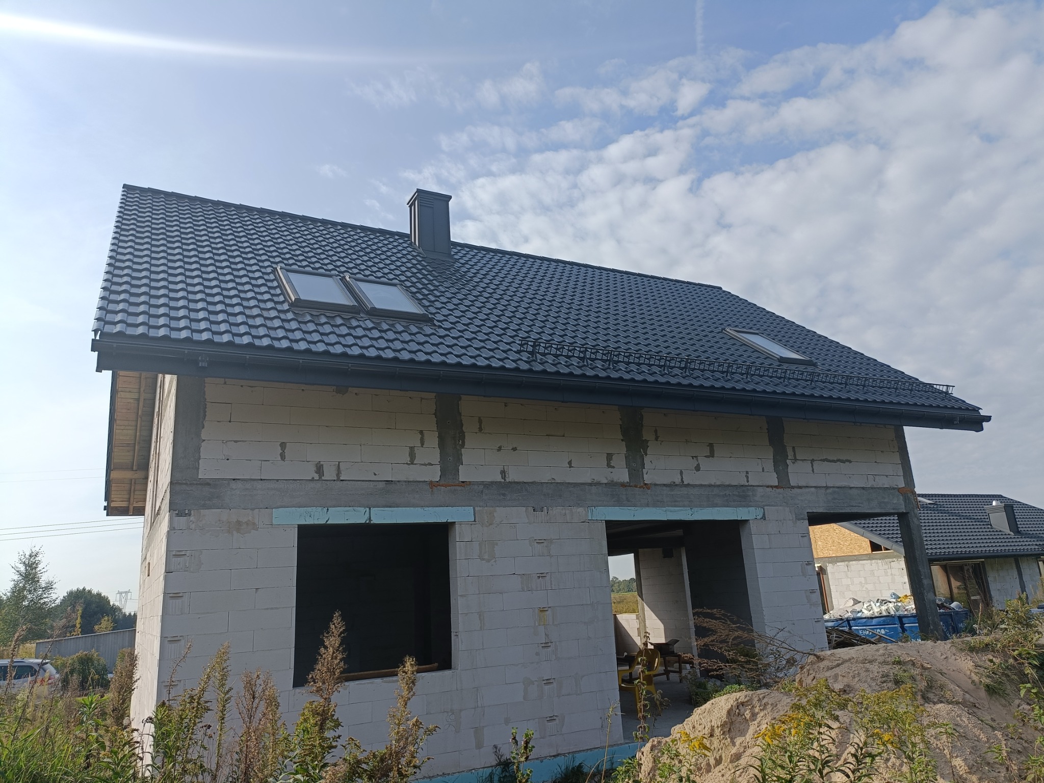 Dom w trakcie budowy z ciemnoszarą dachówką, widoczne okna dachowe i komin, ściany z betonu komórkowego w stanie surowym otwarte, nieotynkowane.