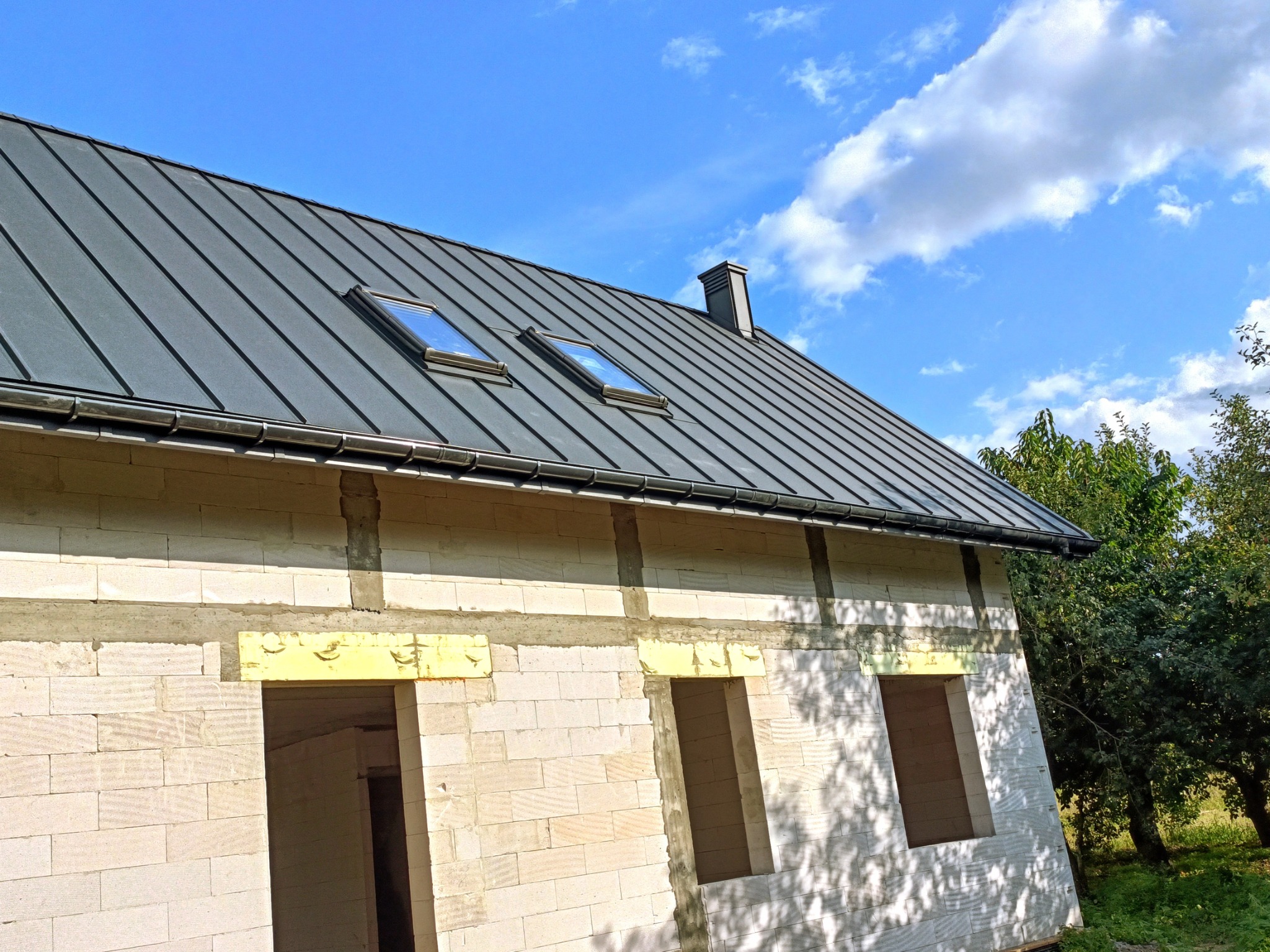 Nowoczesny, grafitowy dach z dwoma oknami dachowymi na surowym budynku z białego bloczka, widok z dołu na błękitne niebo z chmurami.