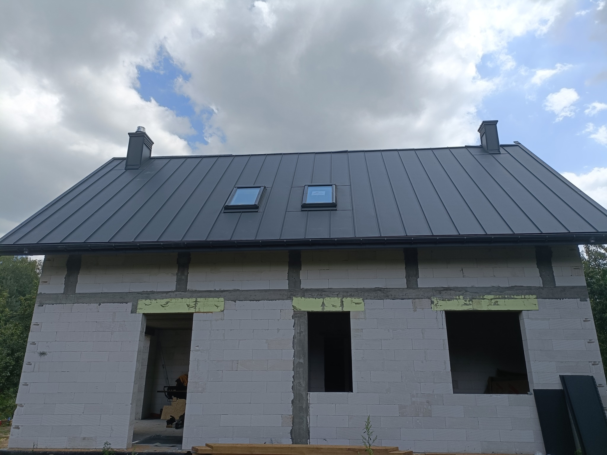 Surowy stan budynku z szarego bloczka z zamontowanym, szarym dachem z blachy na rąbek stojący i dwoma oknami dachowymi, widok z dołu na pochmurne niebo.