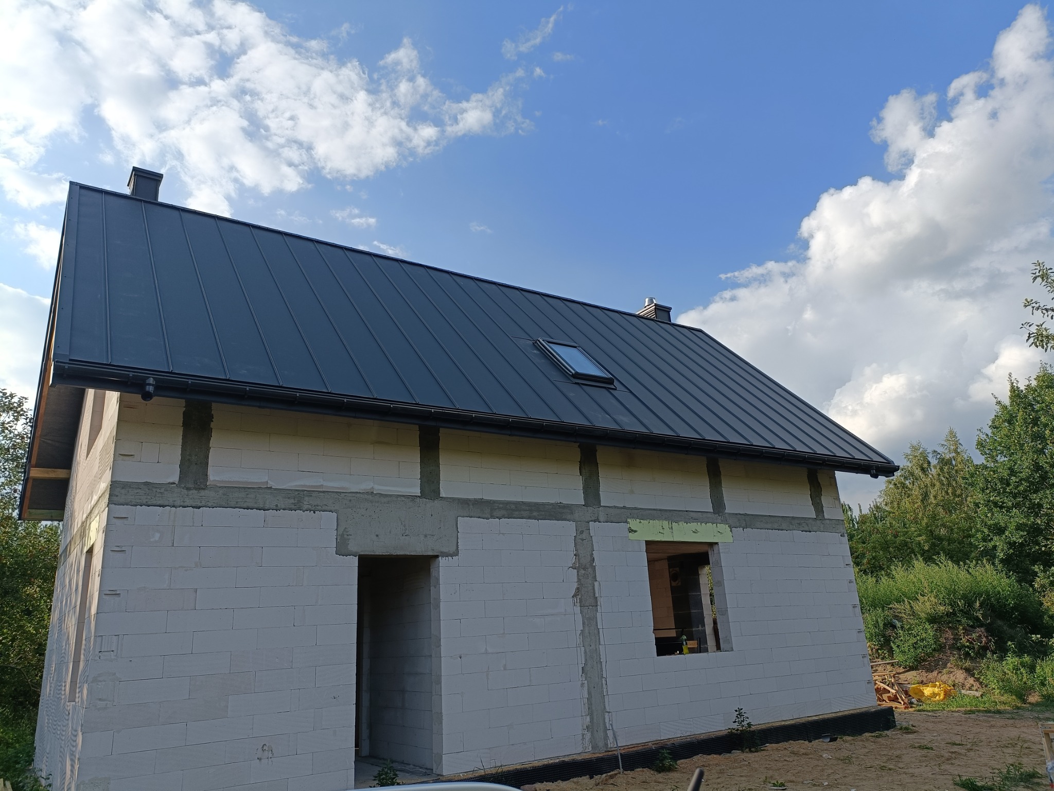 Niedokończony dom z szarego gazobetonu, z zamontowanym, ciemnoszarym dachem z blachy z oknem dachowym i orynnowaniem, na tle błękitnego nieba z obłokami.