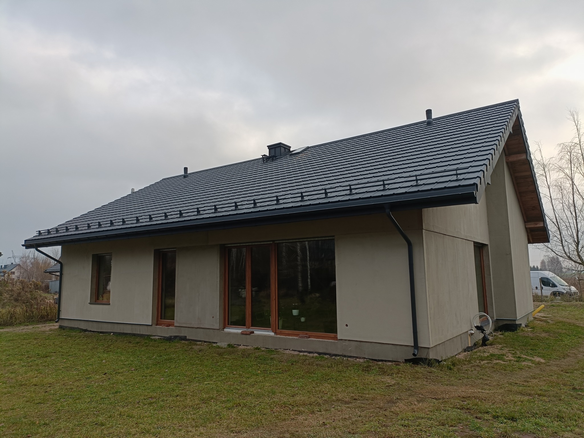 Nowoczesny dom jednorodzinny z ciemnoszarą dachówką ceramiczną i orynnowaniem w kolorze dachu, widok z boku na tle pochmurnego nieba.