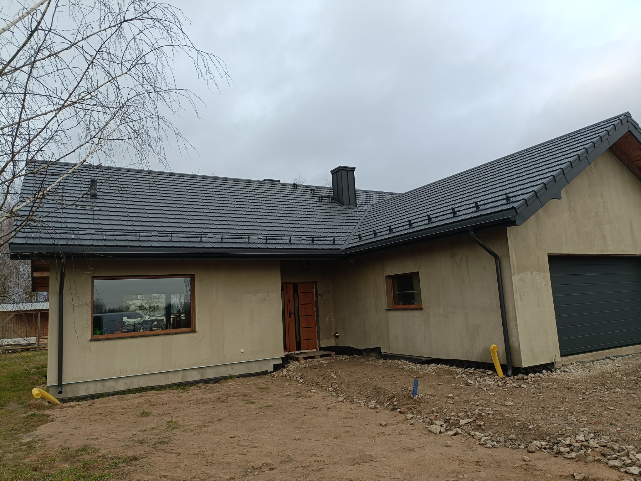 Dom jednorodzinny w trakcie wykończenia z ciemnoszarą dachówką ceramiczną, widoczne rynny, obróbki blacharskie komina i niezabudowane otoczenie.