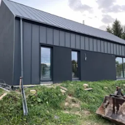 Nowoczesny dom jednorodzinny z grafitową elewacją i blachą trapezową na dachu, widoczne okna z pakietami szybowymi, teren wokół budynku w trakcie prac wykończeniowych, ziemia rozkopana, widoczny...