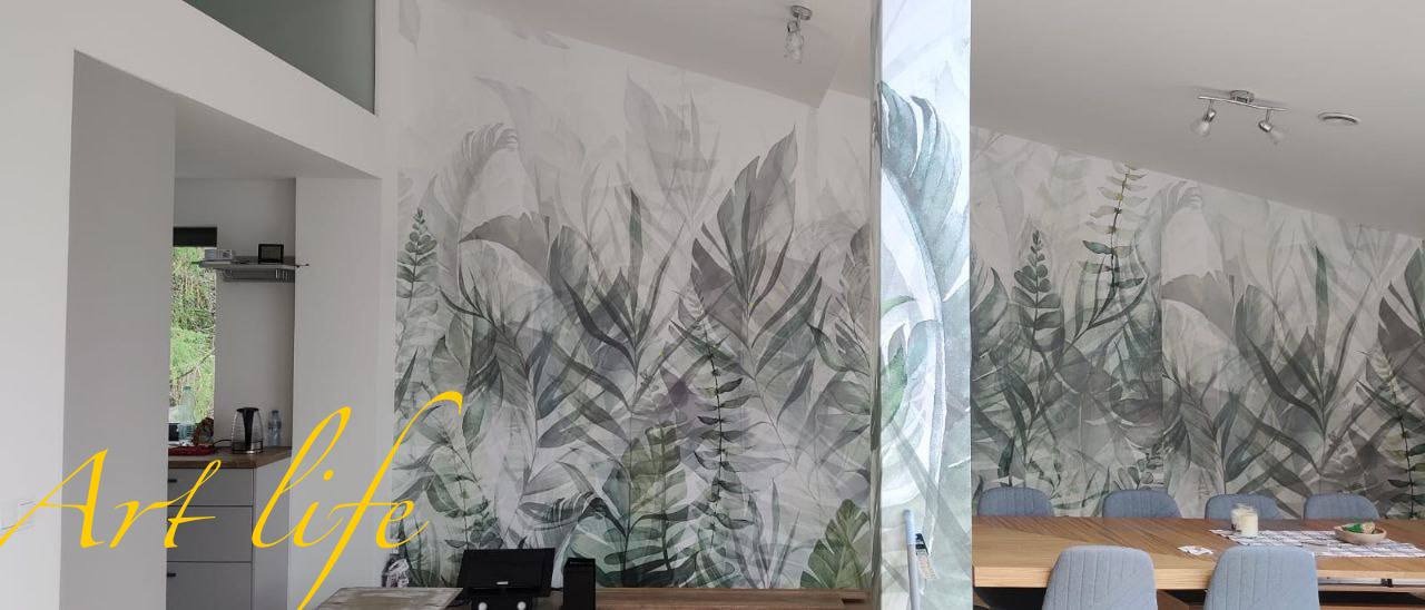 Jasne wnętrze z fototapetą w motywy roślinne, stół z krzesłami w tle, widok na kuchnię z oknem w oddali.