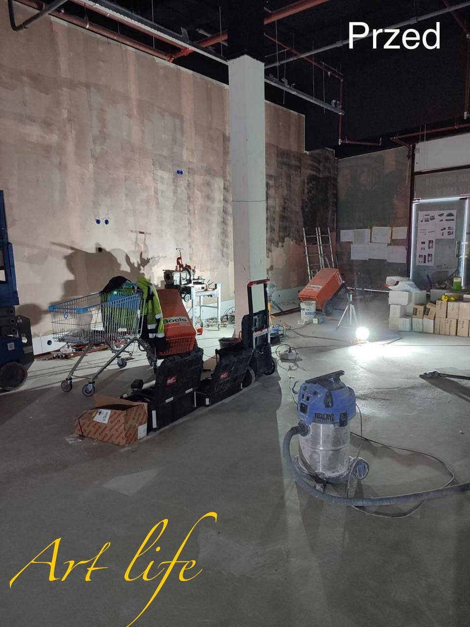 Wnętrze w trakcie prac przygotowawczych: surowe ściany, podłoga pokryta kurzem, widoczne narzędzia, wózek sklepowy z odzieżą roboczą, odkurzacz przemysłowy, oznaczenie 'Przed' w prawym górnym rogu...