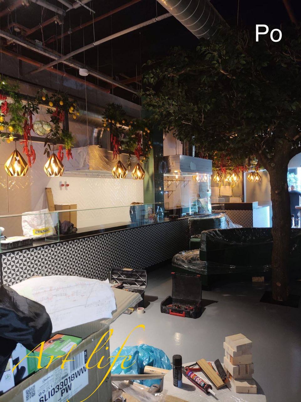 Wnętrze lokalu w trakcie prac wykończeniowych: widoczne narzędzia, materiały budowlane na podłodze, częściowo zakryte meble, dekoracyjne lampy wiszące nad blatem z czarno-białą mozaiką, sztuczne...