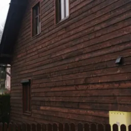 Ciemnobrązowa, drewniana elewacja domu z widocznymi oknami, wentylacją i skrzynką gazową za niskim, drewnianym płotem.