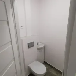 Mała, biała łazienka z podwieszaną toaletą, umywalką z szafką i szarą podłogą z płytek, widok z perspektywy drzwi.