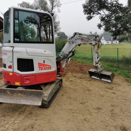 ASBUD - Biało-czerwona mini koparka Takeuchi TB325R na nierównym, piaszczystym terenie z widocznym wykopem, w tle ogrodzenie z siatki i domy jednorodzinne w oddali, pochmurny dzień.