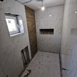 ASBUD - Wnętrze łazienki w trakcie wykańczania, widoczne płytki imitujące marmur na ścianach, fragment ściany wyłożony płytkami drewnopodobnymi, okno, wnęka w ścianie oraz podłoga zabezpieczona folią...
