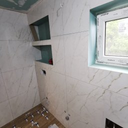 ASBUD - Wnętrze łazienki w trakcie wykańczania w Raba Wyżna: ułożone płytki imitujące drewno na podłodze z użyciem klinów, marmurowe płytki na ścianach z wnękami, okno PCV z widokiem na zieleń.