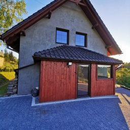 ASBUD - Szary dom z drewnianą dobudówką, czarną dachówką i niebieską kostką brukową na podjeździe, widok z dołu w słoneczny dzień.