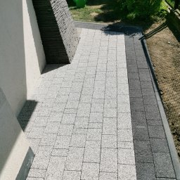 ASBUD - Ułożona kostka brukowa w dwóch odcieniach szarości przylegająca do ściany budynku z dekoracyjnym elementem z kamienia łupanego; w tle zielona konewka i krzew.