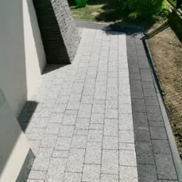 Ułożona kostka brukowa w dwóch odcieniach szarości przylegająca do ściany budynku z dekoracyjnym elementem z kamienia łupanego; w tle zielona konewka i krzew.