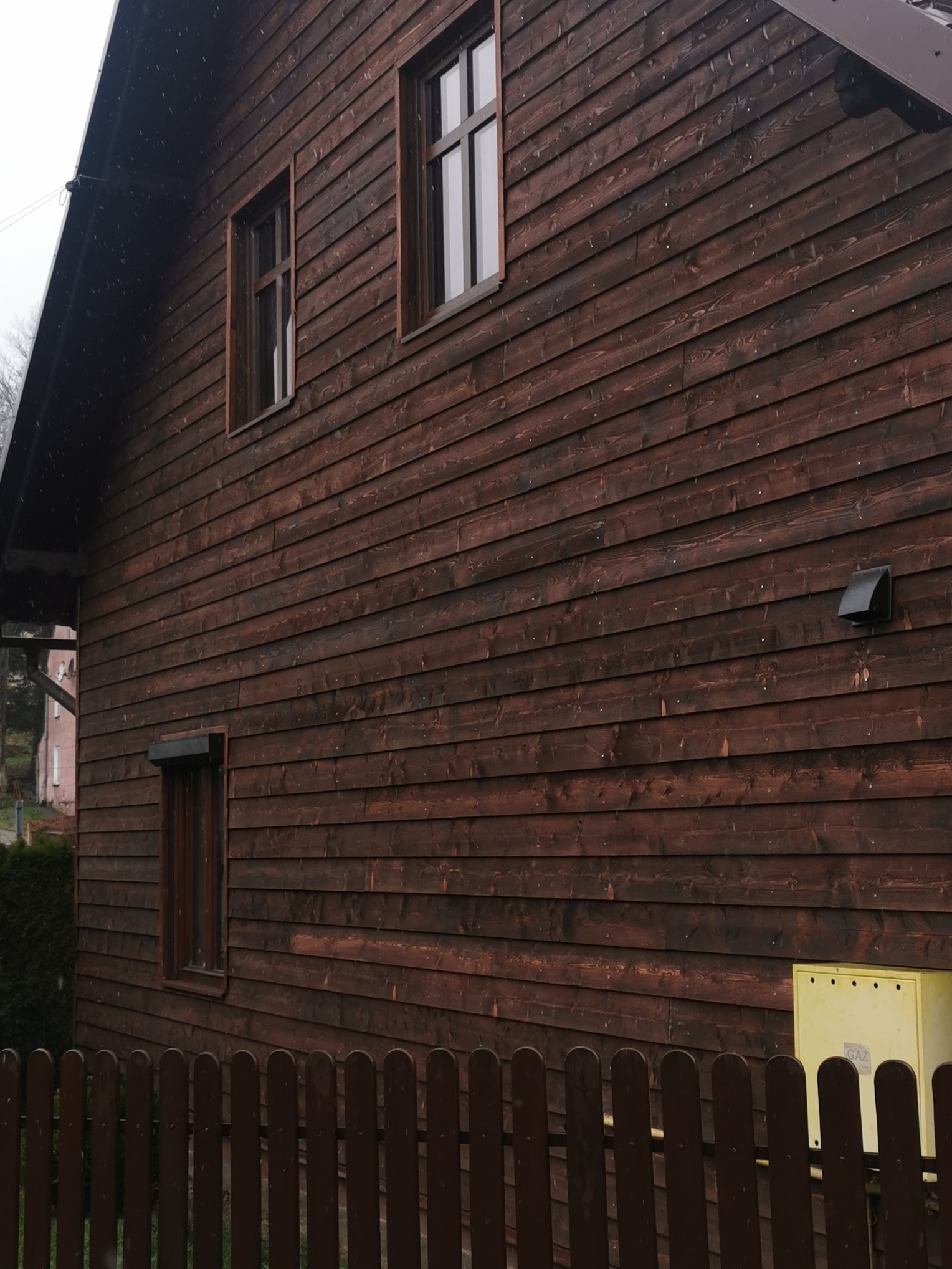 Ciemnobrązowa, drewniana elewacja domu z widocznymi oknami, wentylacją i skrzynką gazową za niskim, drewnianym płotem.