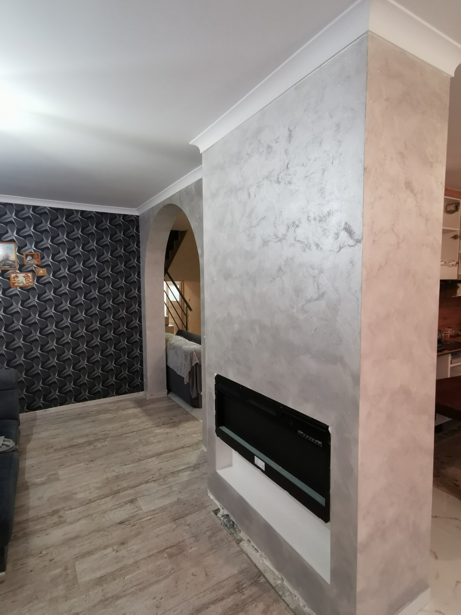 Ściana dekoracyjna z efektem perłowym w odcieniach szarości i różu, z wbudowanym czarnym kominkiem elektrycznym, widok na salon z wzorzystą tapetą i łukowatym przejściem.