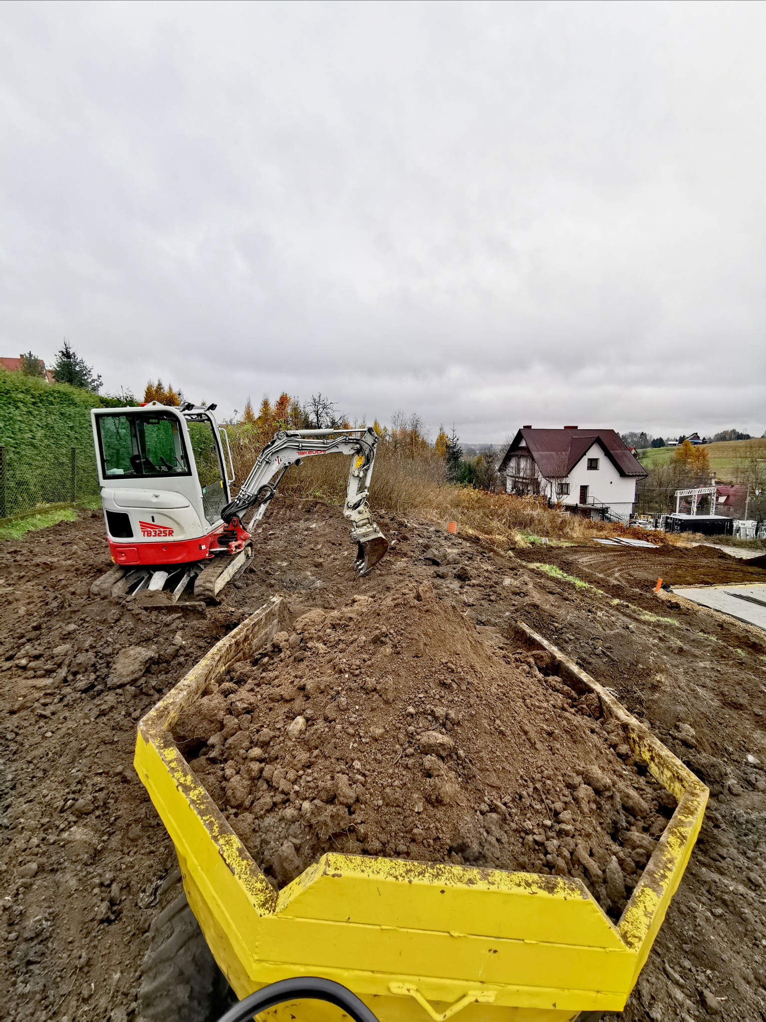 Mini koparka Takeuchi TB325R na placu budowy, z żółtą wywrotką wypełnioną ziemią na pierwszym planie, w tle dom w trakcie budowy i pochmurne niebo.