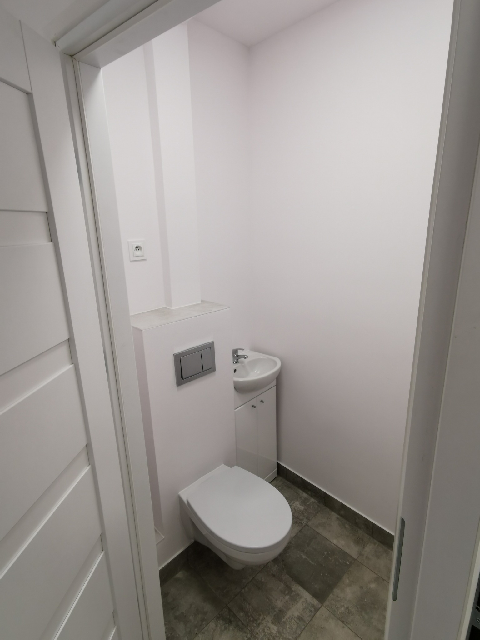 Mała, biała łazienka z podwieszaną toaletą, umywalką z szafką i szarą podłogą z płytek, widok z perspektywy drzwi.