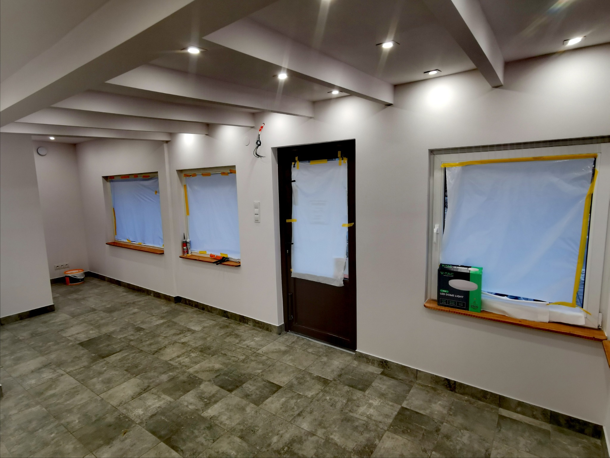 Przygotowane do odświeżenia pomieszczenie z zabezpieczonymi oknami i drzwiami folią malarską, widoczny parkiet imitujący kamień, puszka farby i listwa oświetleniowa LED na parapecie okna.