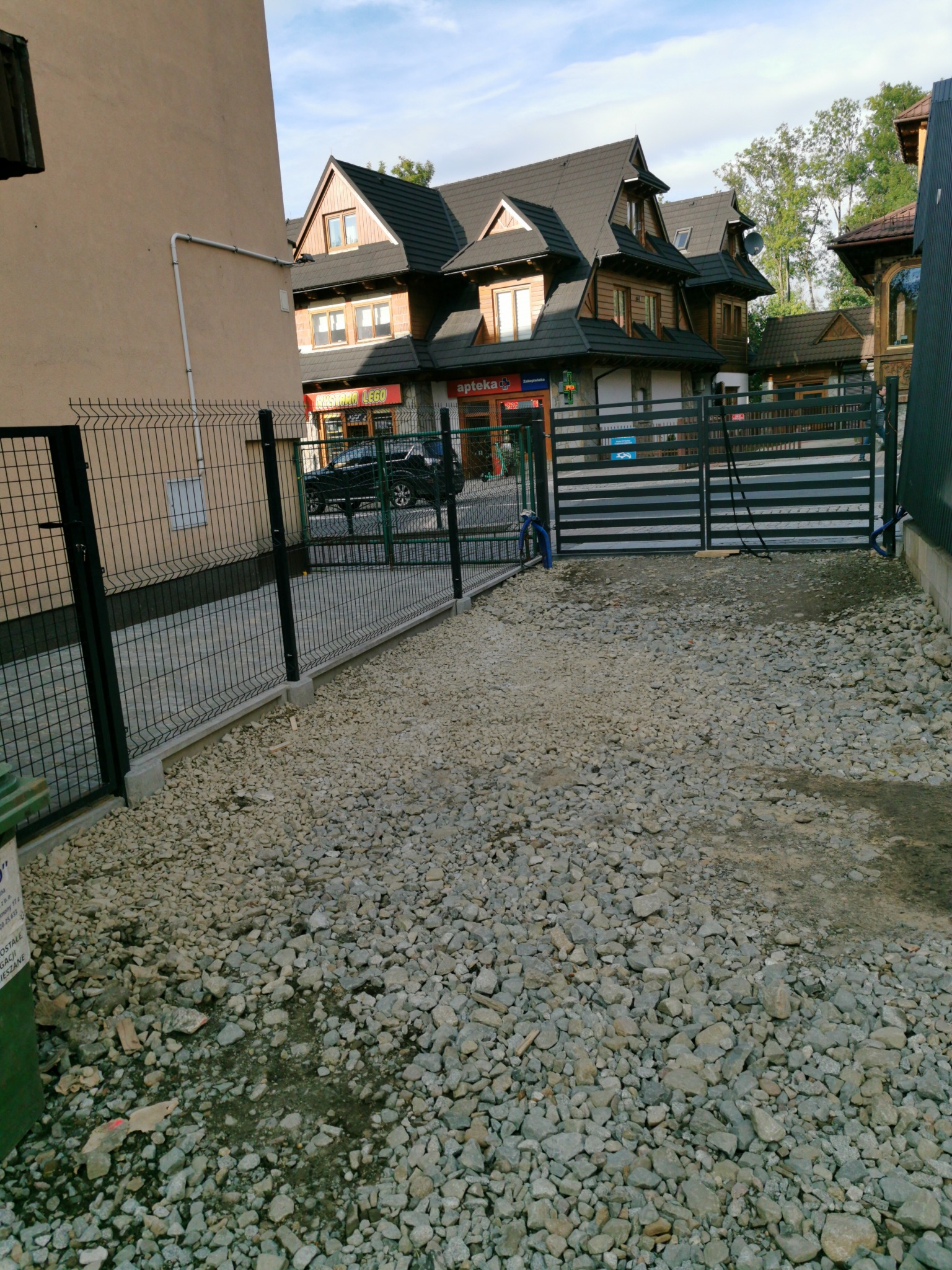 Nowo postawione ogrodzenie panelowe z podmurówką, teren wysypany grysem, w tle budynek z drewnianymi elementami architektonicznymi.