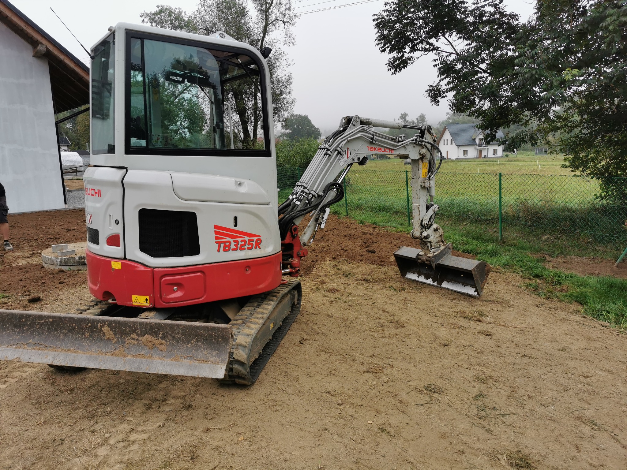 Biało-czerwona mini koparka Takeuchi TB325R na nierównym, piaszczystym terenie z widocznym wykopem, w tle ogrodzenie z siatki i domy jednorodzinne w oddali, pochmurny dzień.