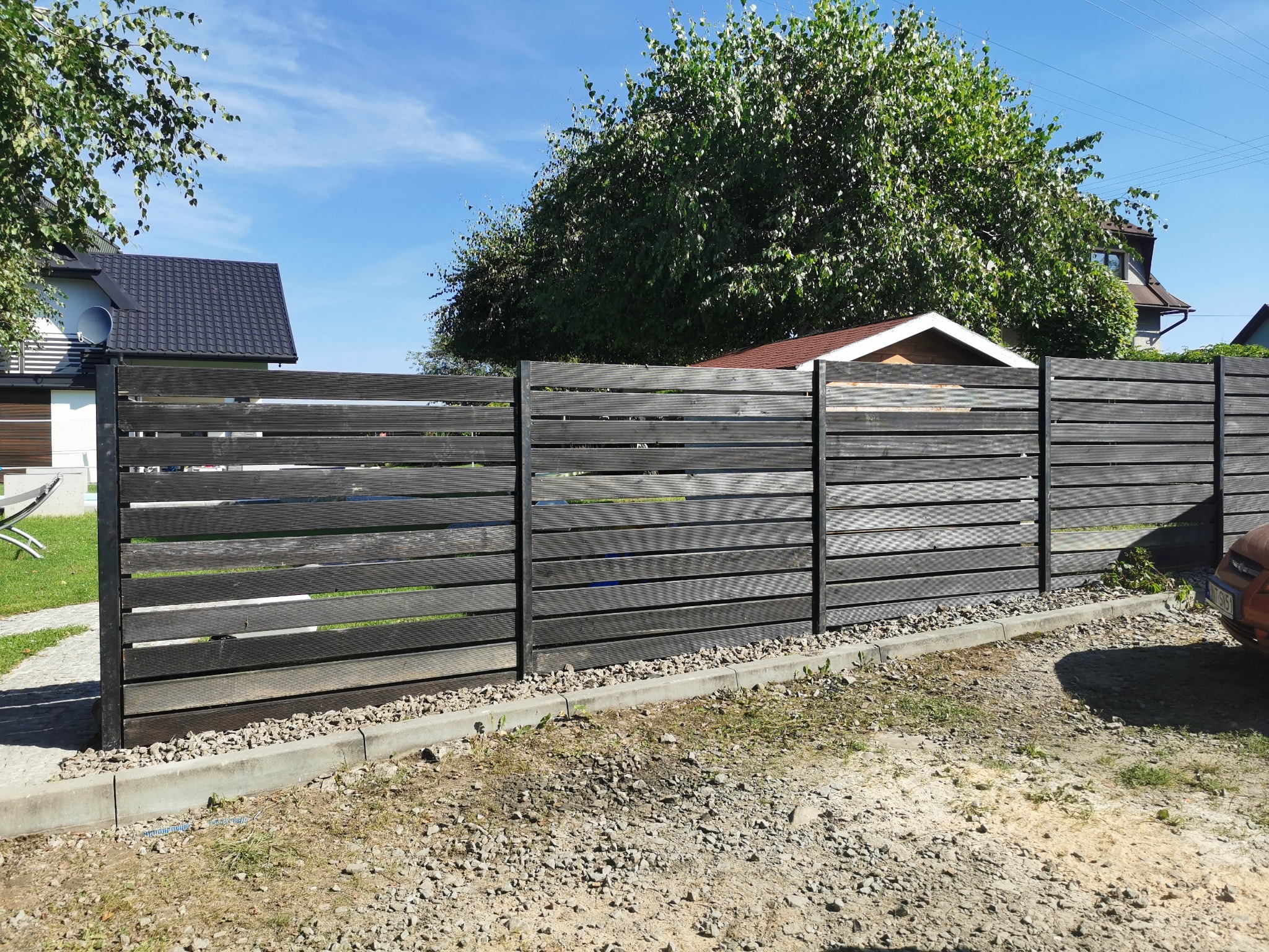 Nowoczesne ogrodzenie panelowe z poziomych desek w kolorze grafitowym, z czarnymi metalowymi słupkami, oddzielające posesję, z widocznym fragmentem domu i zieleni w tle.