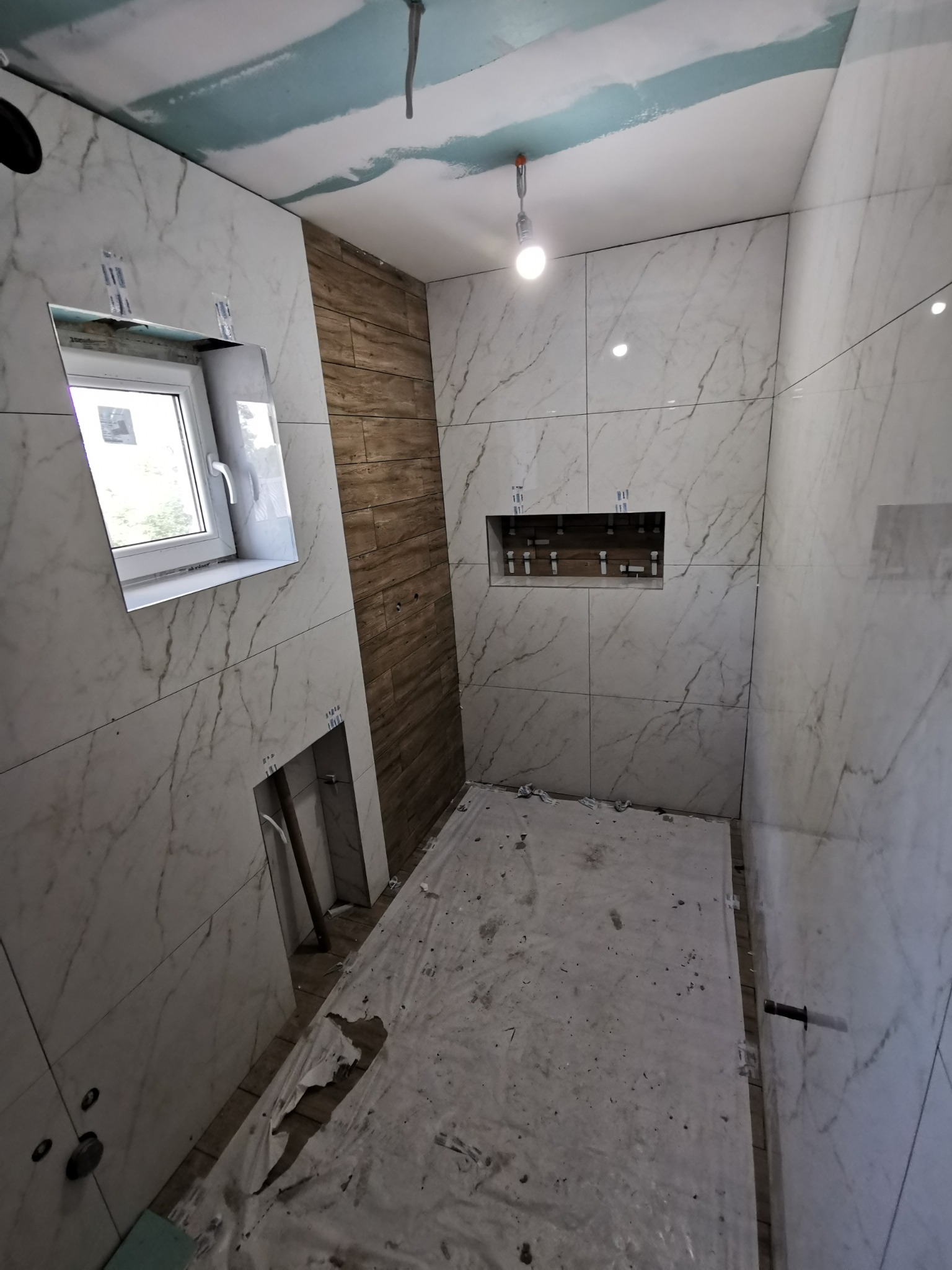 Wnętrze łazienki w trakcie wykańczania, widoczne płytki imitujące marmur na ścianach, fragment ściany wyłożony płytkami drewnopodobnymi, okno, wnęka w ścianie oraz podłoga zabezpieczona folią...