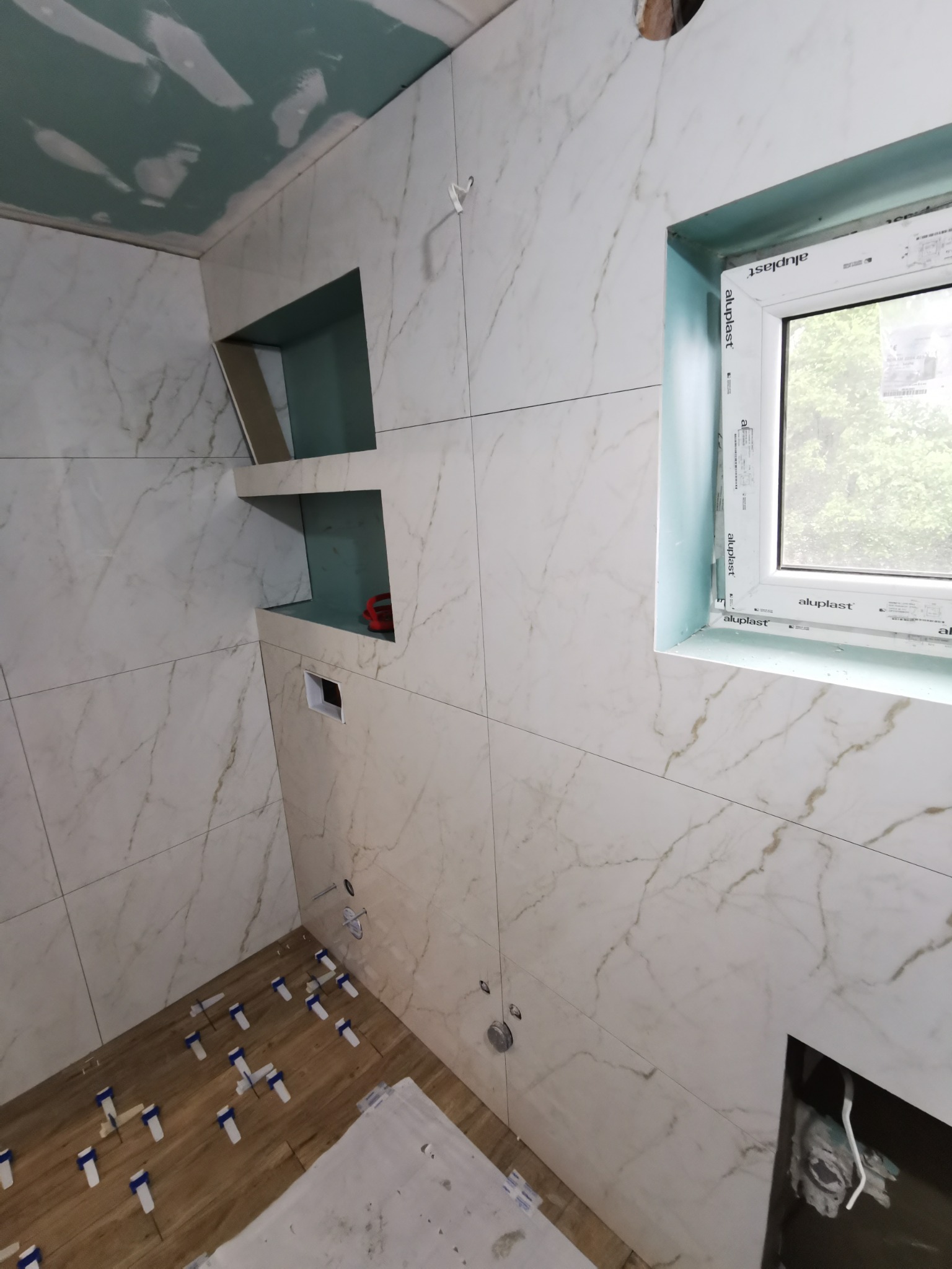 Wnętrze łazienki w trakcie wykańczania w Raba Wyżna: ułożone płytki imitujące drewno na podłodze z użyciem klinów, marmurowe płytki na ścianach z wnękami, okno PCV z widokiem na zieleń.
