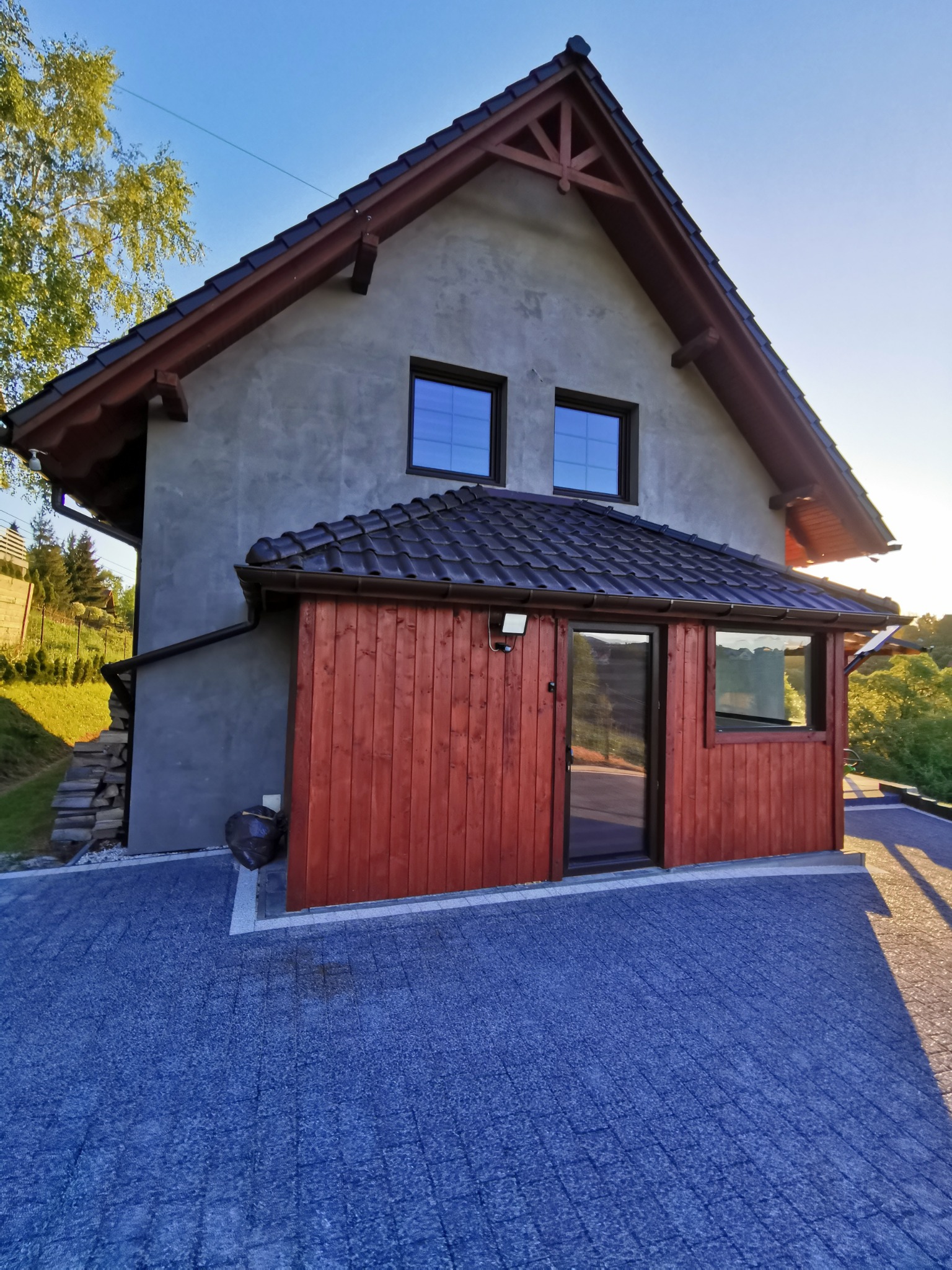 Szary dom z drewnianą dobudówką, czarną dachówką i niebieską kostką brukową na podjeździe, widok z dołu w słoneczny dzień.