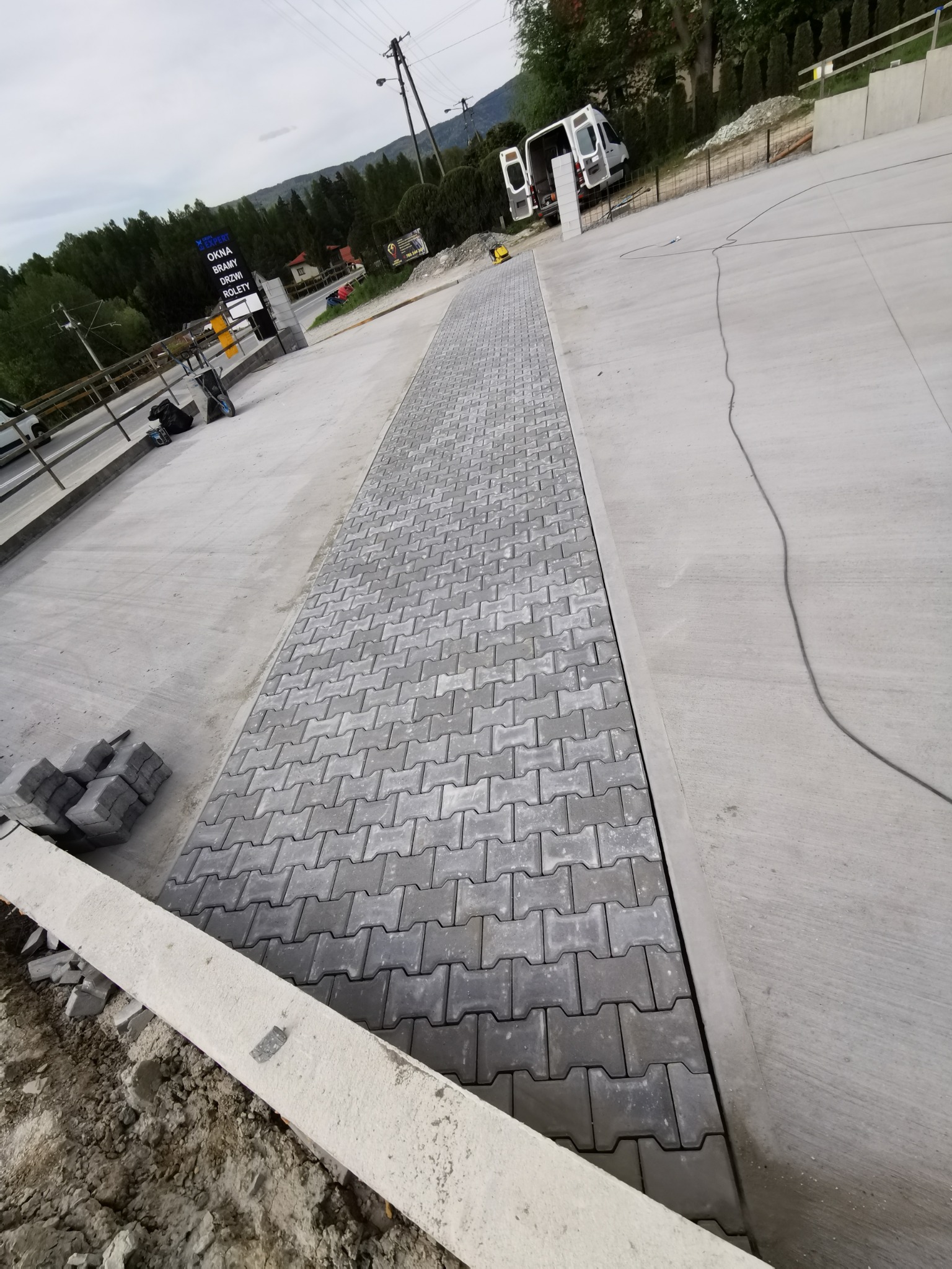 Układanie szarej kostki brukowej na betonowej powierzchni, widoczne zapasy kostki i narzędzia, częściowo ukończony ciąg komunikacyjny.