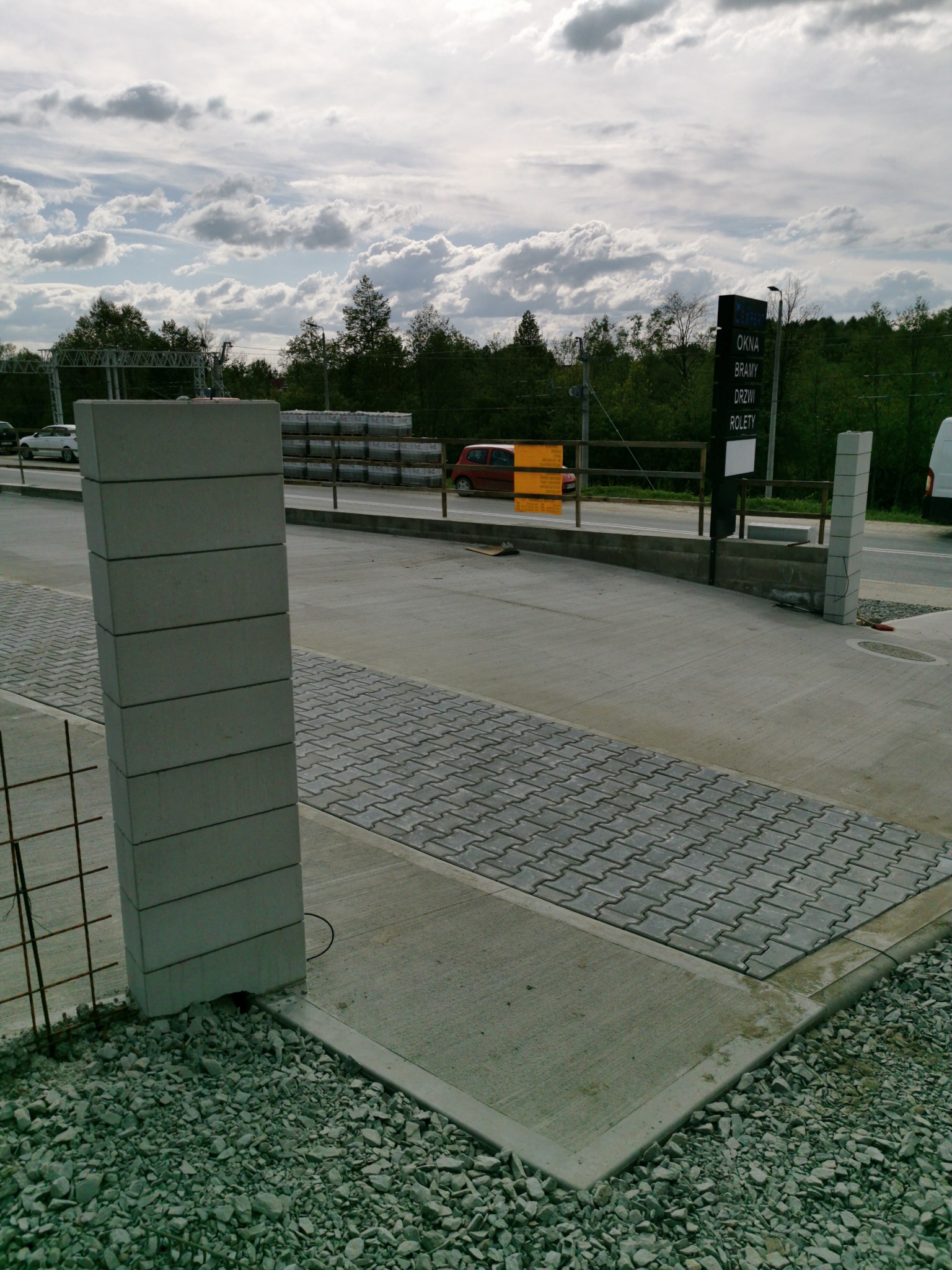 Wznoszenie słupka z betonowych bloczków na posesji, widoczne ułożone kostki brukowe i podsypka z kamieni, w tle budynek z reklamą okien i bram.