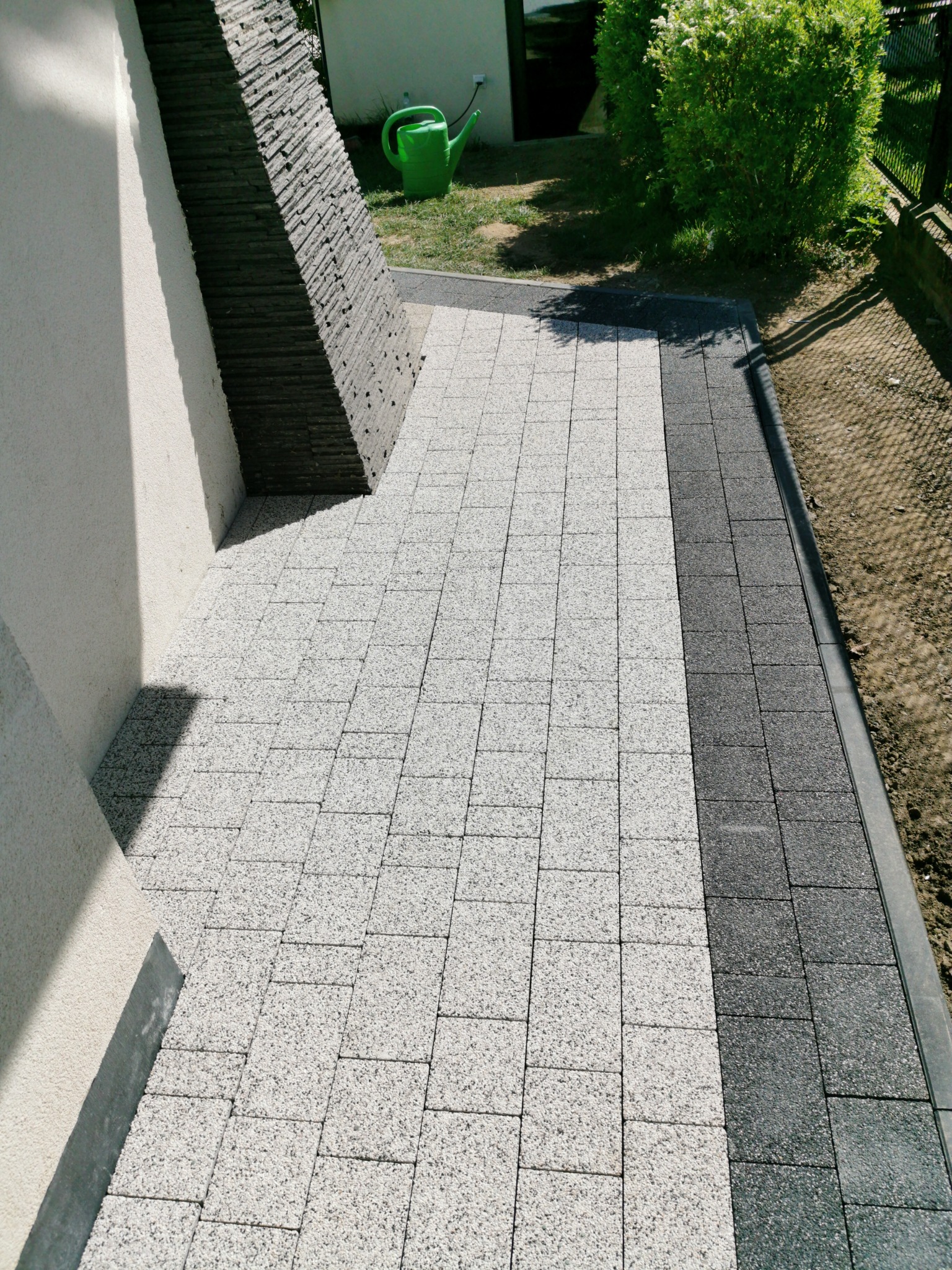 Ułożona kostka brukowa w dwóch odcieniach szarości przylegająca do ściany budynku z dekoracyjnym elementem z kamienia łupanego; w tle zielona konewka i krzew.